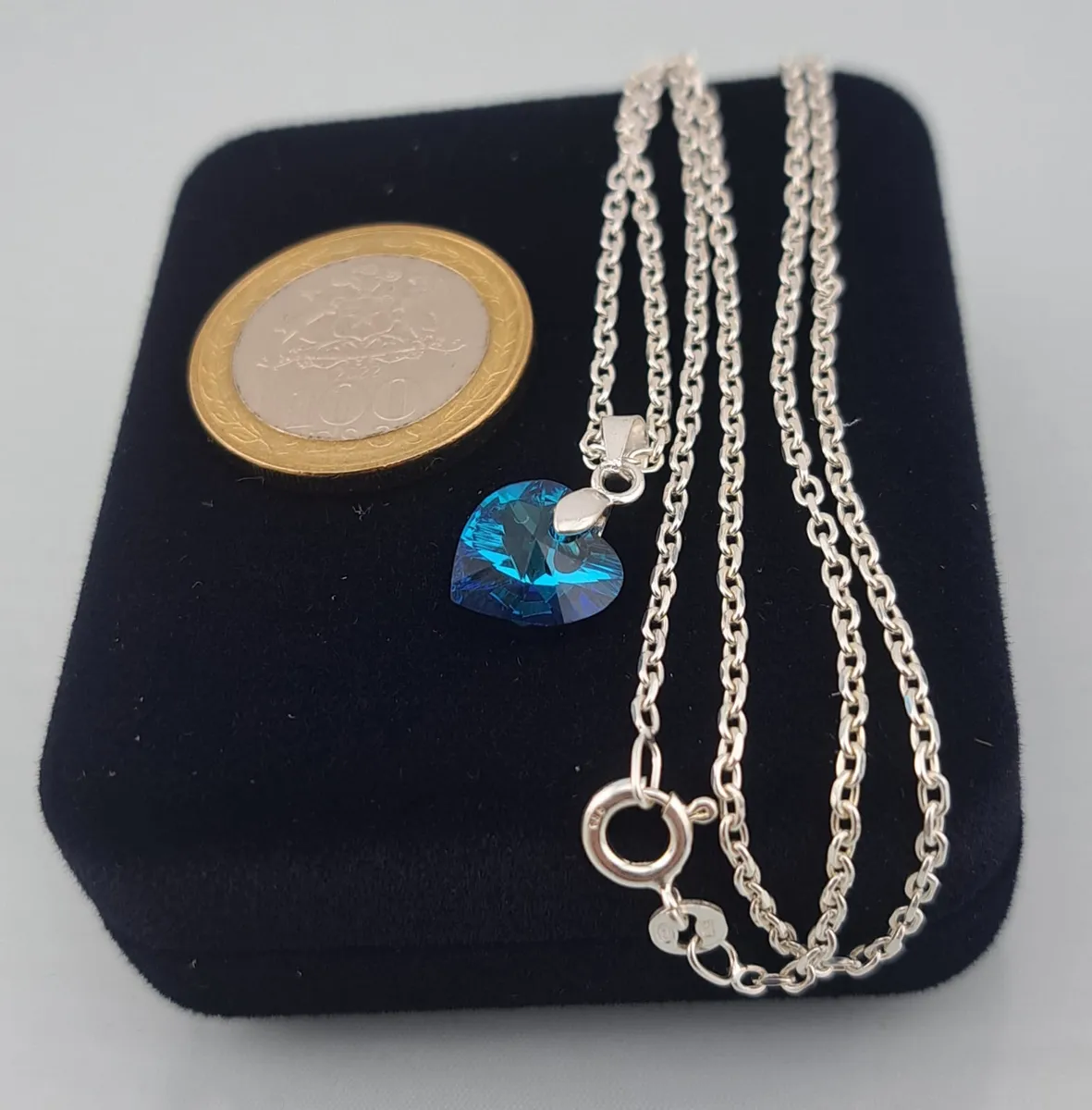 Dije de corazón 10mm de cristal azul visto de perfil con cadena de plata fina y una moneda de 100 pesos chilenos
