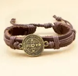 Detalle de pulsera de cuero marrón con amuleto de San Benito sobre superficie neutra.